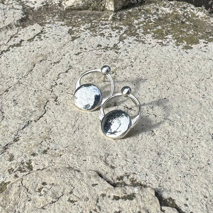 A pair of small silver  hoop earrings with flat circular disc charms and small bead details, photographed on a textured stone surface.