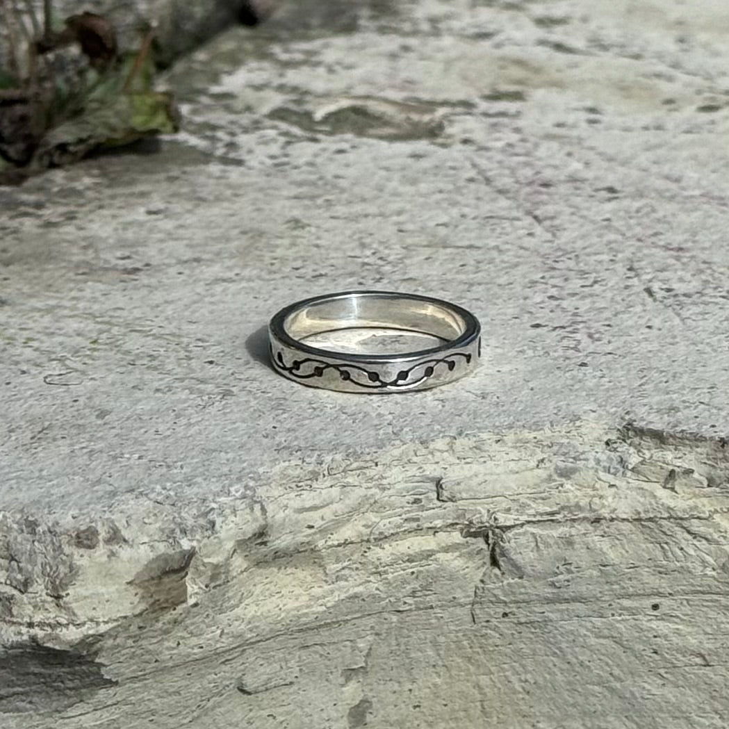 A silver band ring with a black wavy crop circle pattern, resting on a textured gray stone surface.
