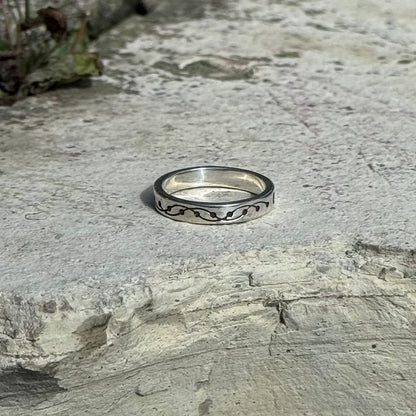 A silver band ring with a black wavy crop circle pattern, resting on a textured gray stone surface.