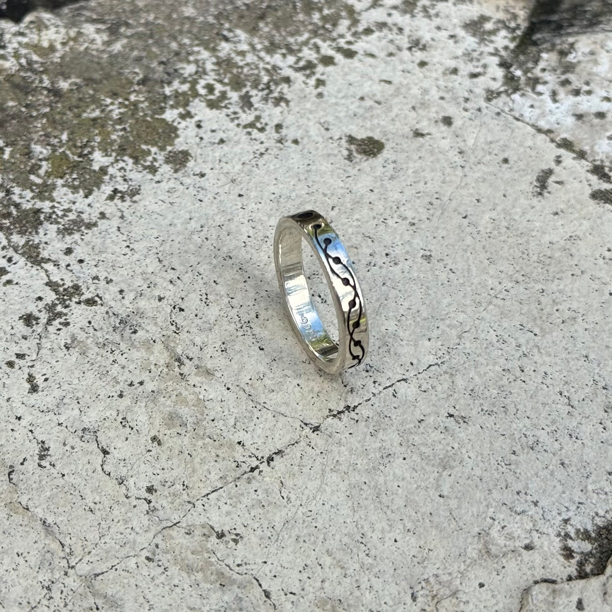 A polished silver ring with a black engraved crop circle design, resting on a textured gray concrete surface.