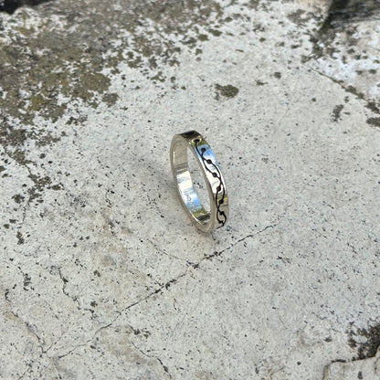 A polished silver ring with a black engraved crop circle design, resting on a textured gray concrete surface.