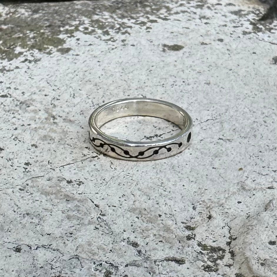 A sterling silver band ring with a black wavy, crop circle inlay pattern, resting on a textured, light gray stone surface.