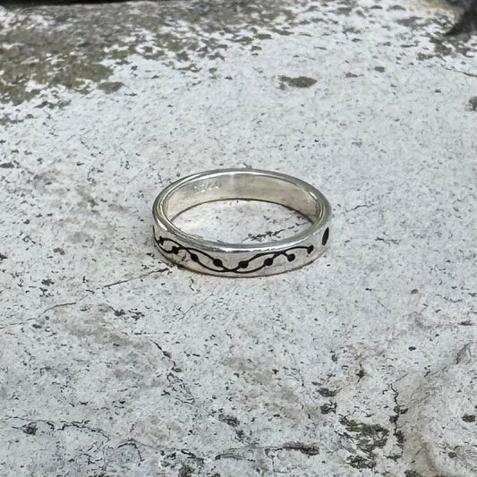 A sterling silver band ring with a black wavy, crop circle inlay pattern, resting on a textured, light gray stone surface.
