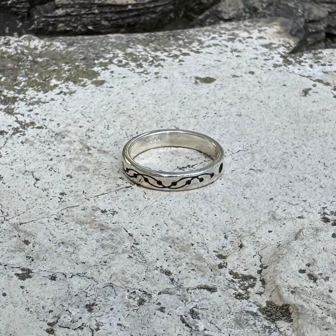 A silver band ring with a black wavy crop circle motif, resting on a textured gray stone surface.