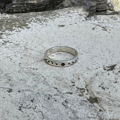 A sterling silver band ring with a decorative crop circle wavy pattern and small black dots, resting on a textured gray stone surface.